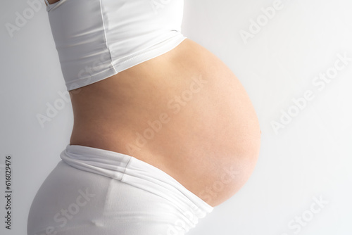 Middle part of unrecognizable standing woman with very round pregnant baby bump. Side view. White background. Bright shot.