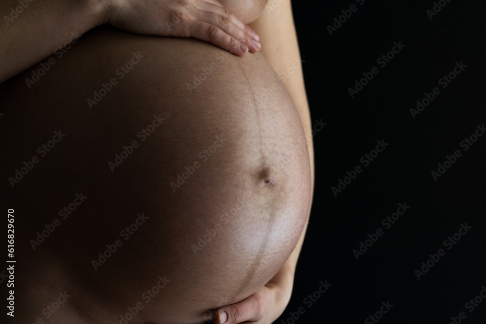Closeup of round pregnant baby bump held by hands of unrecognizable standing naked woman. Final month of pregnancy - week 36. Side angle view. Black background. Closeup of round pregnant baby bump held by hands of unrecognizable standing naked woman. Final month of pregnancy - week 36. Side angle view. Black background.
