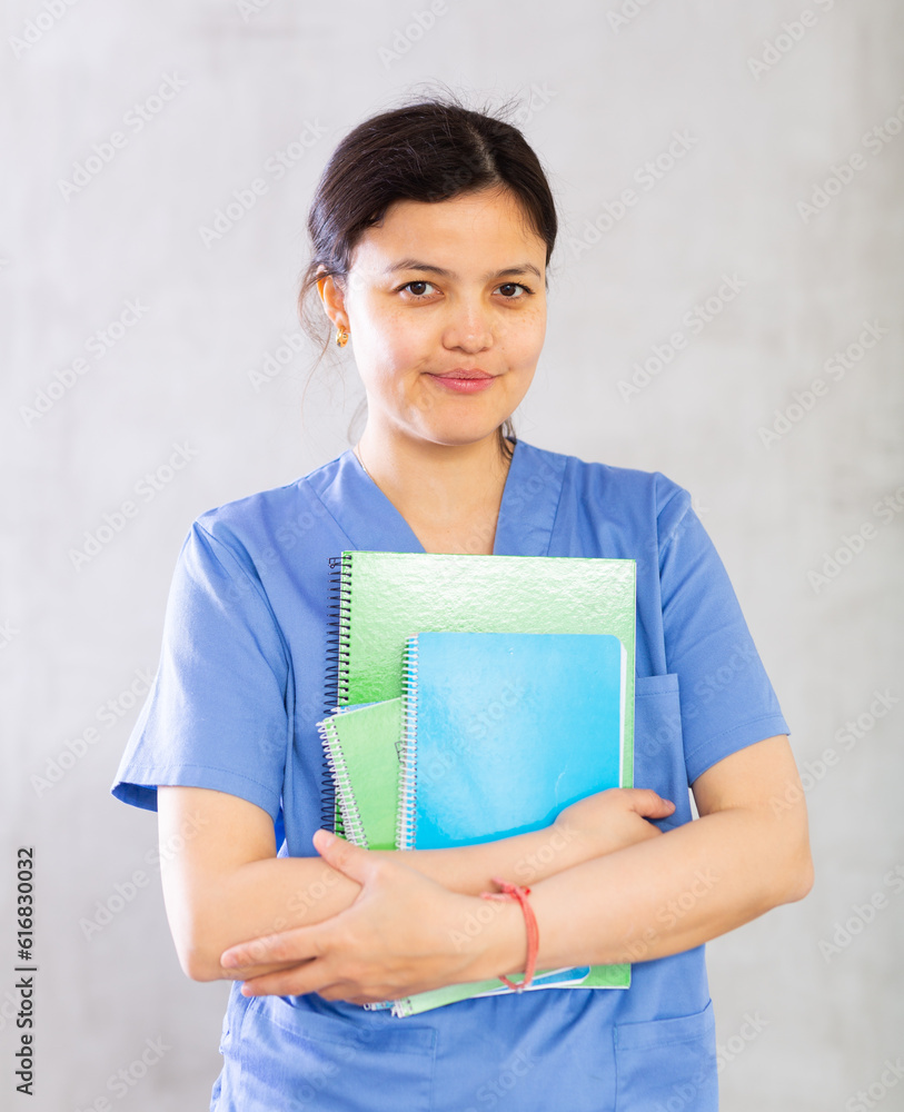 Obraz premium Portrait of smiling young female doctor in blue uniform standing with notebooks indoors on gray background..