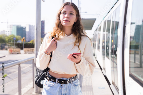 Confident girl with a backpack on her back, walking along a tram stop, holds a mobile phone in her hands photo