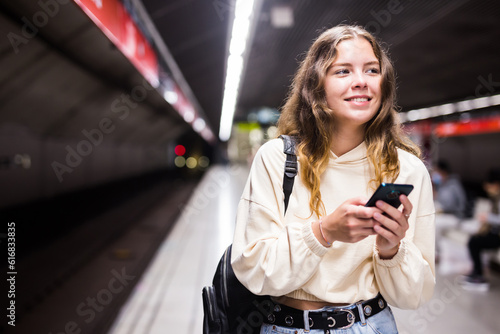 Positive girl with a backpack standing on the platform of a metro station, texting on her mobile phone with friends, ..waiting for the train