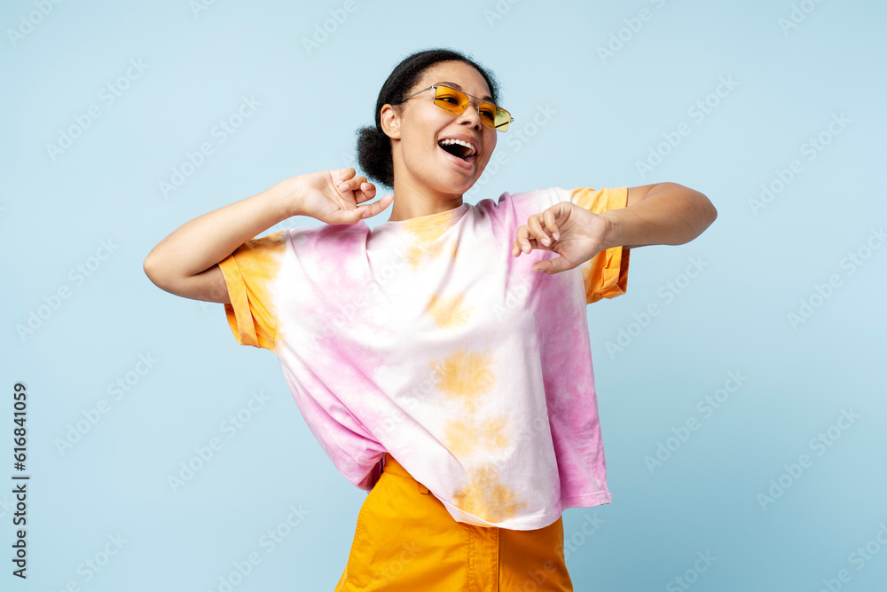 Smiling African American woman dancing wearing trendy t shirt and eyeglasses isolated on blue background. Happy stylish fashion model having fun posing for picture in studio Smiling African American woman dancing wearing trendy t shirt and eyeglasses isolated on blue background. Happy stylish fashion model having fun posing for picture in studio