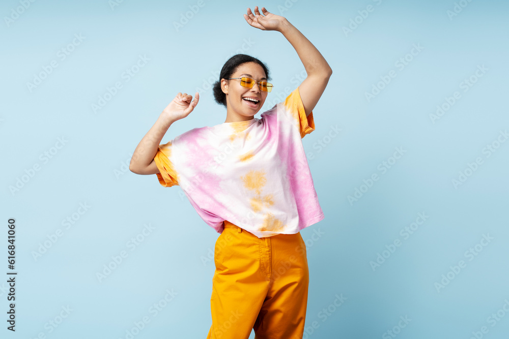 Portrait of beautiful happy African American woman dancing wearing trendy tie dye t shirt isolated on blue background. Smiling stylish fashion model having fun posing for picture in studio Portrait of beautiful happy African American woman dancing wearing trendy tie dye t shirt isolated on blue background. Smiling stylish fashion model having fun posing for picture in studio