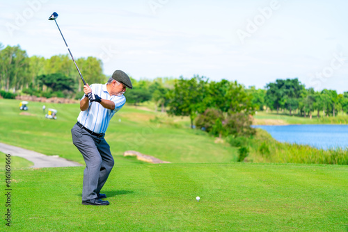 Asian senior man holding golf club hitting golf ball on fairway at country club in sunny day. Healthy retired elderly man golfer enjoy outdoor activity sport golfing at golf course on summer vacation