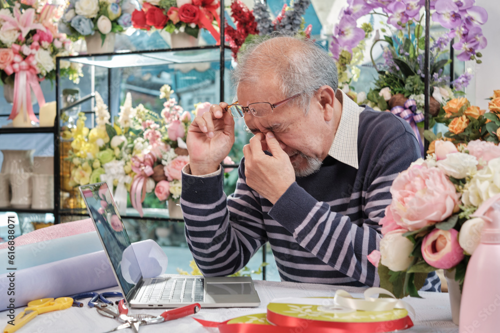 Asian elderly male florist owner is unwell with headaches and health deteriorating, stress from overwork in a flower shop, SME business entrepreneur. Asian elderly male florist owner is unwell with headaches and health deteriorating, stress from overwork in a flower shop, SME business entrepreneur.