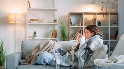 Sick tea. Virus drinking. Sore throat. Shivering woman catching cold suffering influenza holding cup healing grippe sitting wrapped blanket on couch in living room at home.