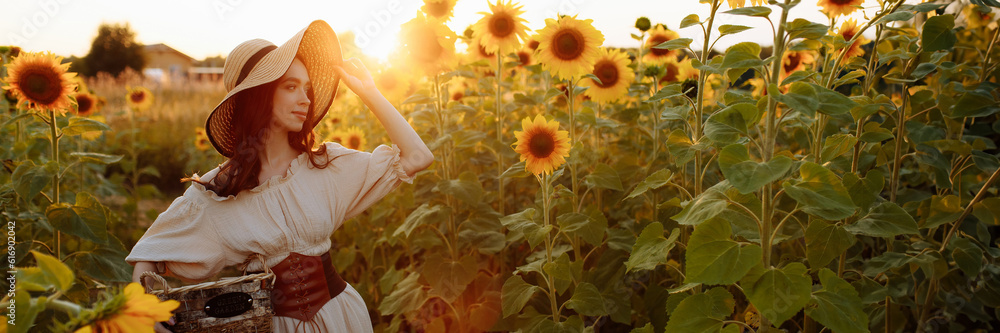 A beautiful young tall slender brunette woman in a beige dress and leather corset stands in a field with blooming yellow sunflowers at sunset. Banner for website header design with copy space.