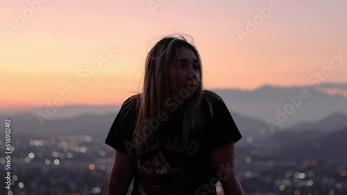 beautiful latina young girl sitting in front of a mountain sunset at mt rubidoux in riverside california with pink and oranges hues and blurred city lights in the background photo