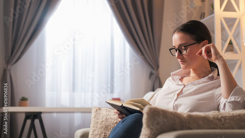 Reading hobby. Home leisure. Weekend literature. Relaxed curious smart woman learning with open book on cozy couch at modern living room interior with free space.