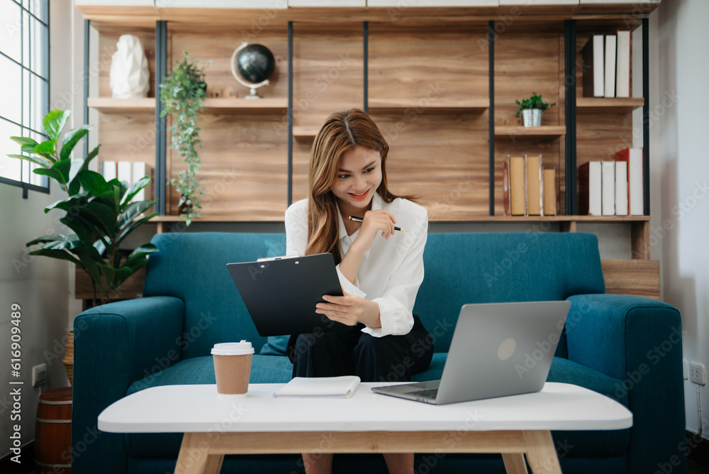 Fototapeta premium Young attractive Asian woman smiling thinking planning writing in notebook, tablet and laptop working from home at office .