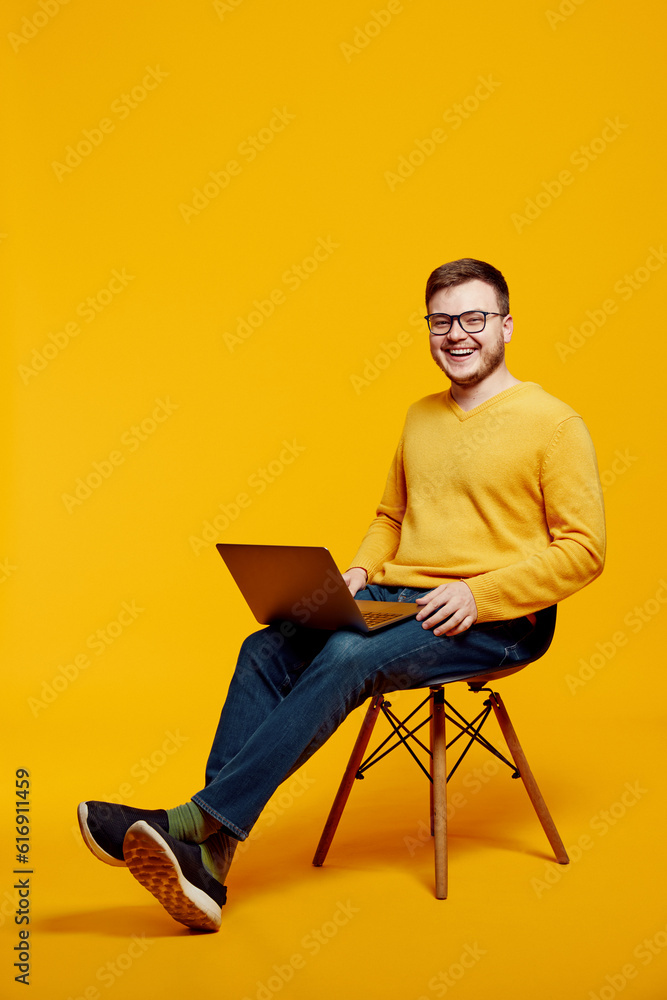 Caucasian cheerful adult man wearing yellow sweater and eyeglasses, using laptop computer while sitting on chair, isolated over orange background Caucasian cheerful adult man wearing yellow sweater and eyeglasses, using laptop computer while sitting on chair, isolated over orange background