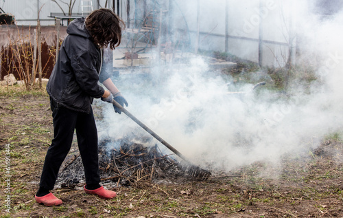 A young woman in a black jacket burns dry grass in the yard