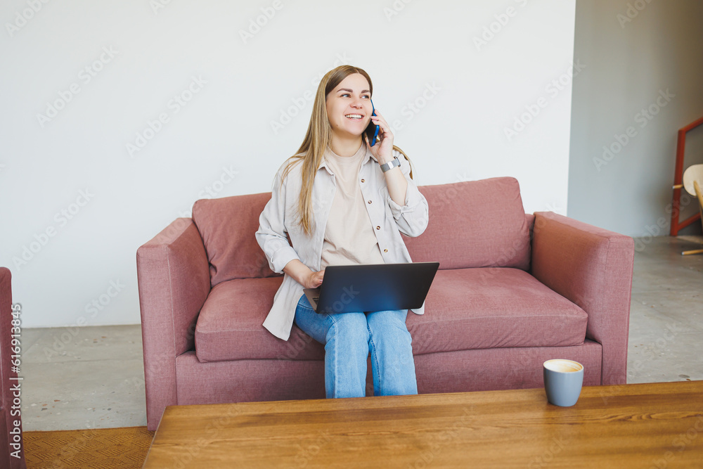 Smiling female freelancer in casual clothes sitting in comfortable sofa in modern cafeteria at table with laptop and phone, talking on mobile phone. Remote work Smiling female freelancer in casual clothes sitting in comfortable sofa in modern cafeteria at table with laptop and phone, talking on mobile phone. Remote work