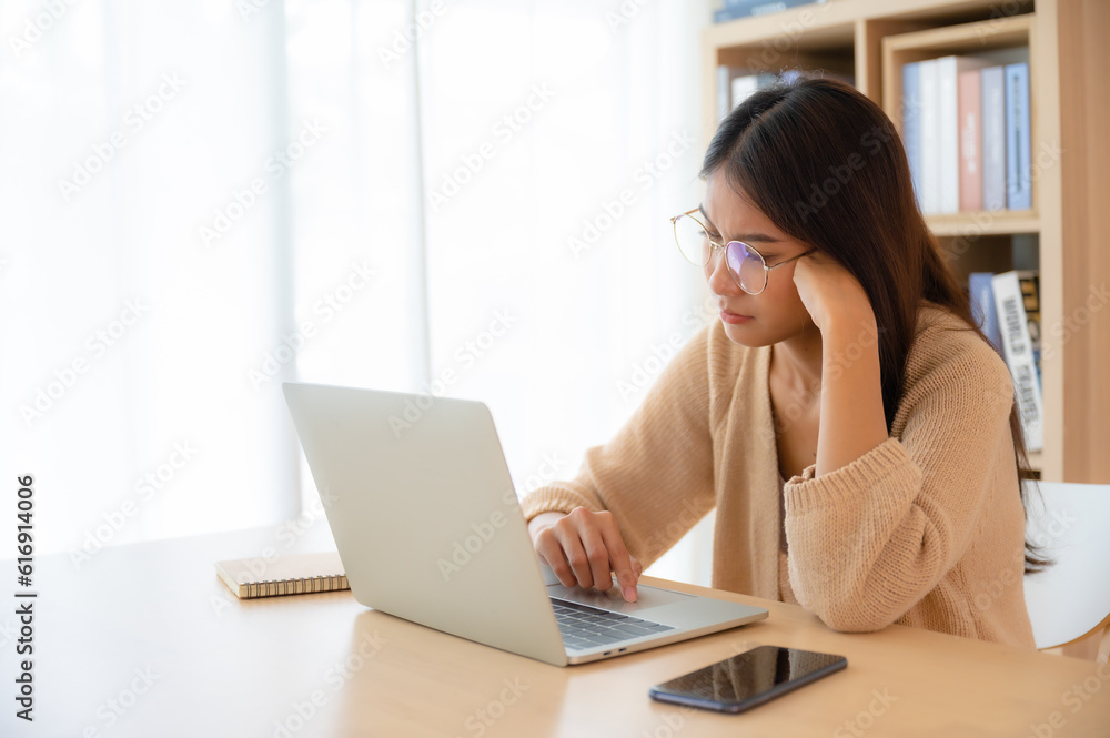 Young Asian Woman Working on Computer Laptop in the Living Room, Experiencing Emotional Turmoil, Serious, Stress, Loneliness, Anxiety, and Despair Young Asian Woman Working on Computer Laptop in the Living Room, Experiencing Emotional Turmoil, Serious, Stress, Loneliness, Anxiety, and Despair