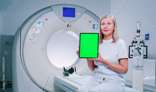 Mature woman is sitting at TC scanner bed. Female at room of MRI. Woman dressed up in white is showing screen of chroma key tablet. Female patient is posing at background of medical equipment. photo