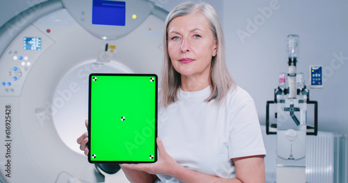 Close-up of woman sitting at TC scanner bed. Mature female at room of MRI. Woman dressed up in white is showing screen of chroma key tablet. Female patient is smiling and posing at camera. photo