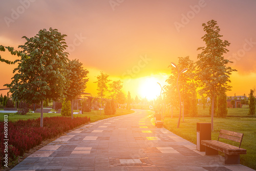 City park in early summer or spring with pavement, lanterns, young green lawn, trees and dramatic cloudy sky on a sunset or sunrise.
