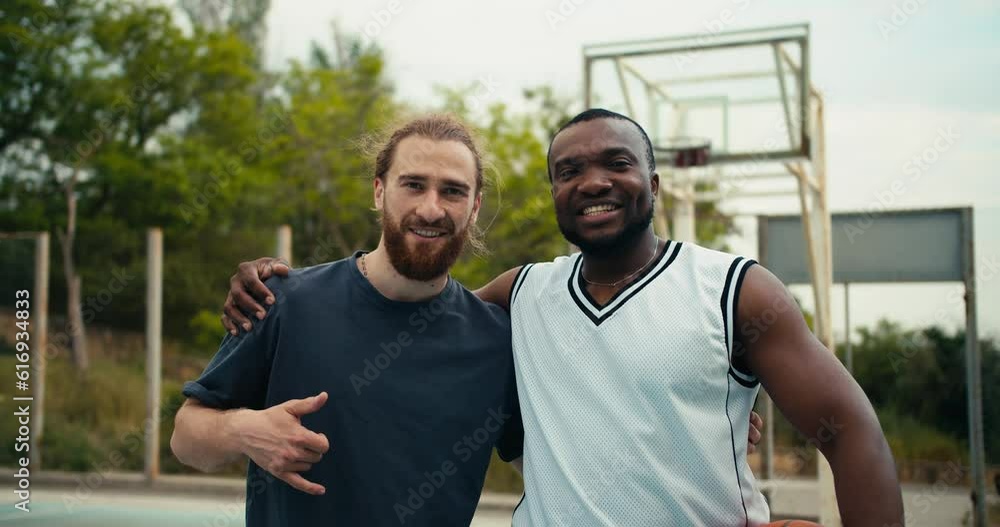 Portrait of two friends, a red-haired man and a black man in a white t-shirt, posing, looking at the camera and smiling on the background of a basketball court Portrait of two friends, a red-haired man and a black man in a white t-shirt, posing, looking at the camera and smiling on the background of a basketball court