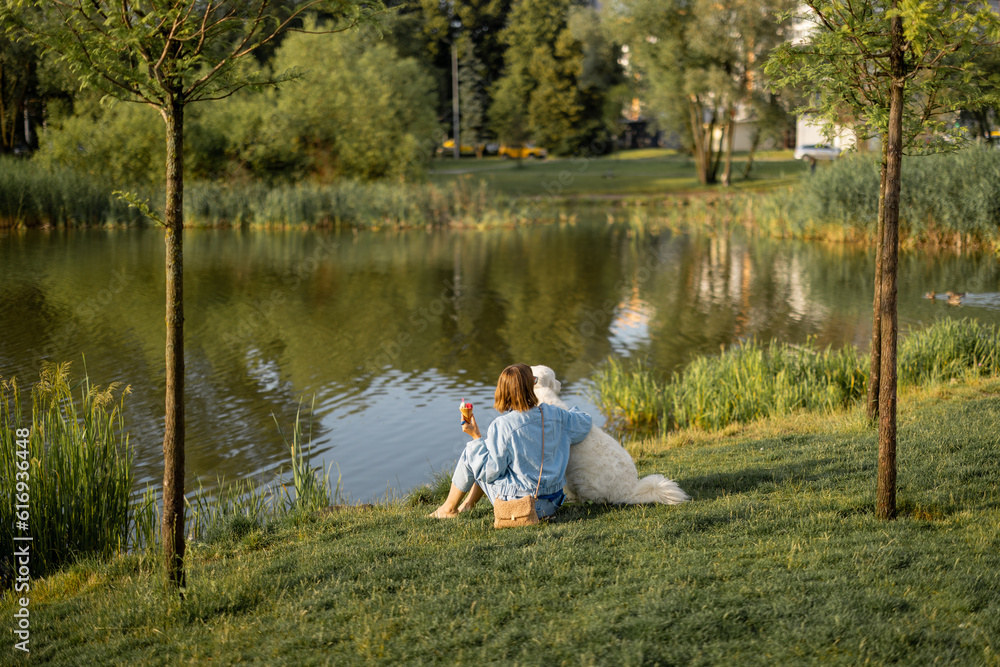 Young woman eats ice cream while sitting with her cute white dog near the lake in park. Wide view. Friendship with pets and spending leisure time together Young woman eats ice cream while sitting with her cute white dog near the lake in park. Wide view. Friendship with pets and spending leisure time together