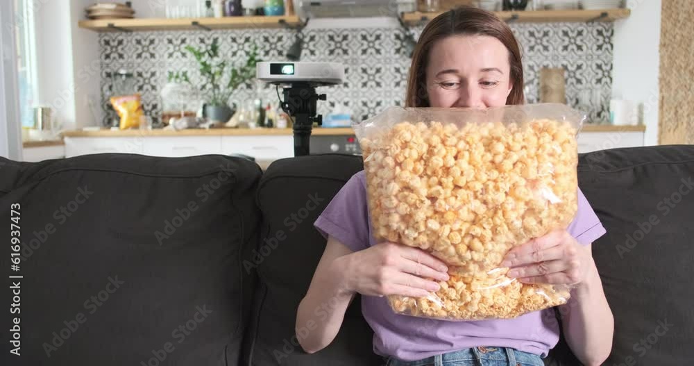 Young woman shakes huge bag popcorn. Happy girl rejoices before watching movie on home video LED projector. Young woman shakes huge bag popcorn. Happy girl rejoices before watching movie on home video LED projector.