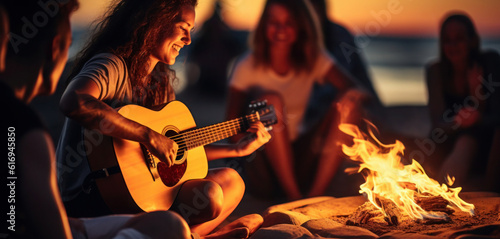 Young people playing guitar and singing songs while having fun in a group while sitting next to a campfire on a beach at night.