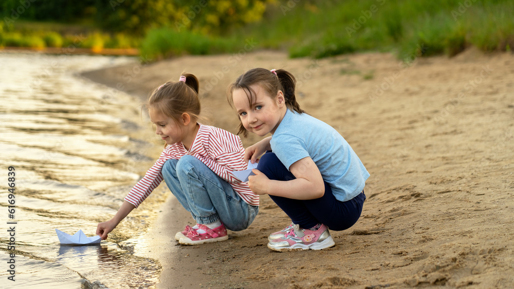 Little girls play with paper boats on river on background of summer park at sunset. Children walk outdoors by water Little girls play with paper boats on river on background of summer park at sunset. Children walk outdoors by water