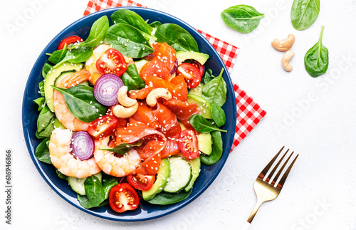 Fresh salad for keto diet with shrimp, salmon, avocado, spinach, cucumber, tomato, cashew nuts, sesame. Low-carbohydrate lunch rich in healthy fats. White table background, top view