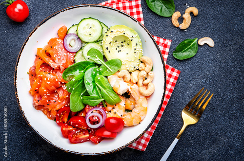 Keto diet salad with salmon, shrimp, avocado, spinach, cucumber, tomato, cashew, sesame. Low-carbohydrate lunch rich in healthy fats. Black table background, top view