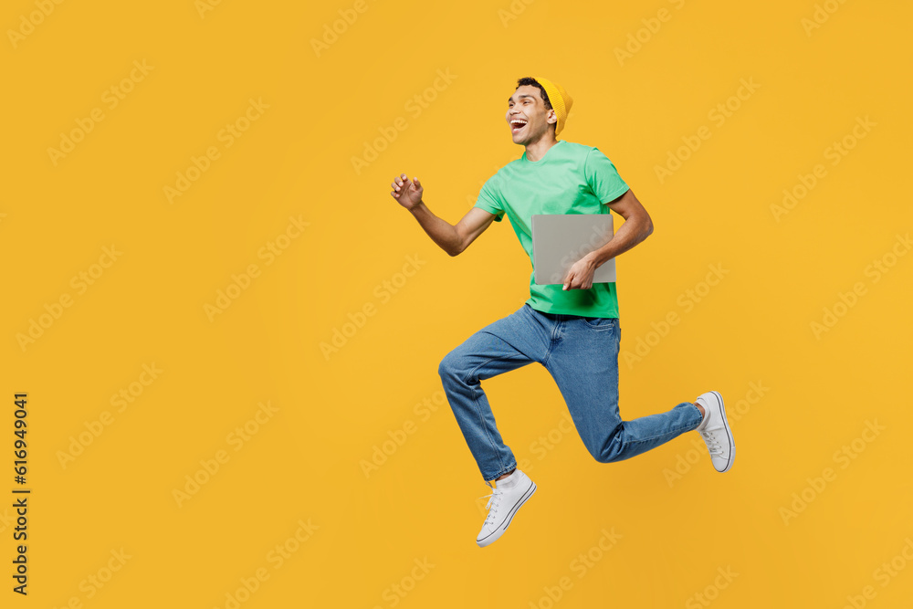 Full body young IT man of African American ethnicity wearing casual clothes green t-shirt hat jump high hold closed laptop pc computer run fast isolated on plain yellow background. Lifestyle concept.