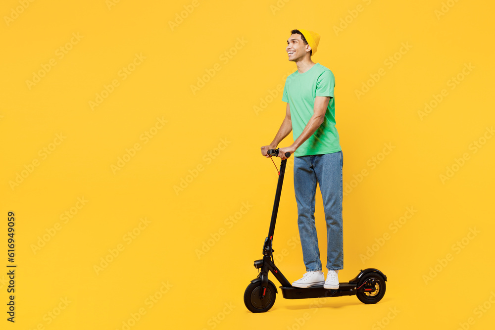Full body side view young man of African American ethnicity he wearing casual clothes green t-shirt hat riding electric scooter isolated on plain yellow background studio portrait. Lifestyle concept. Full body side view young man of African American ethnicity he wearing casual clothes green t-shirt hat riding electric scooter isolated on plain yellow background studio portrait. Lifestyle concept.