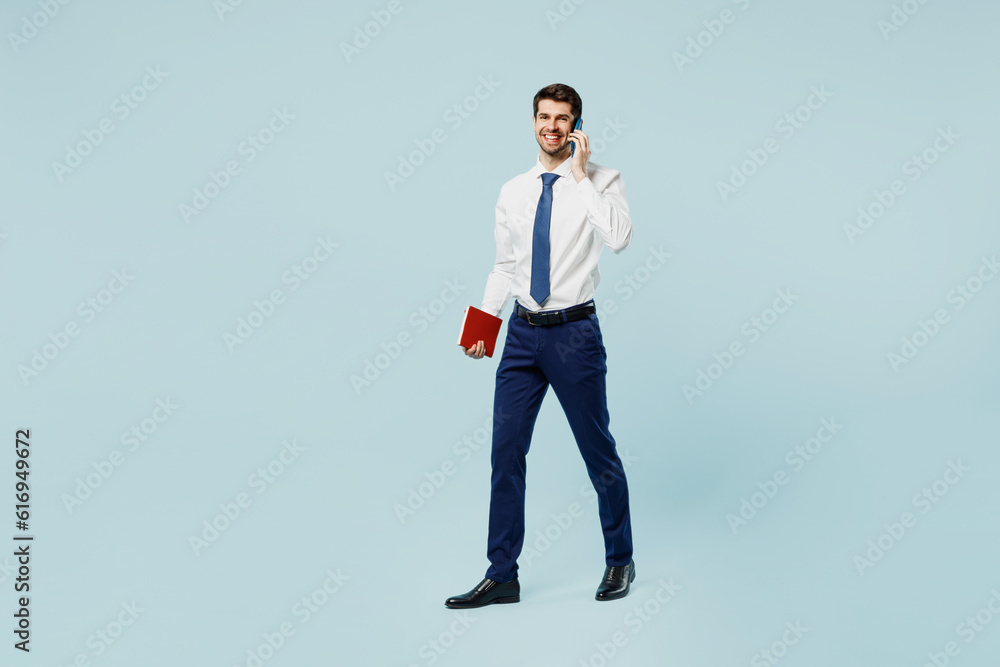 Full body young employee IT business man corporate lawyer wear classic formal shirt tie work in office talk speak on mobile cell phone hold book isolated on plain pastel light blue background studio. Full body young employee IT business man corporate lawyer wear classic formal shirt tie work in office talk speak on mobile cell phone hold book isolated on plain pastel light blue background studio.