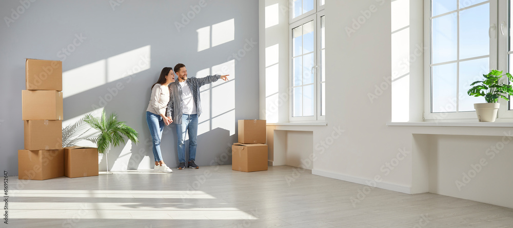 © Studio Romantic - Happy family couple moving to a new house or apartment. Joyful young man and woman homeowners standing in empty living room interior at home and looking through window at view. Banner background