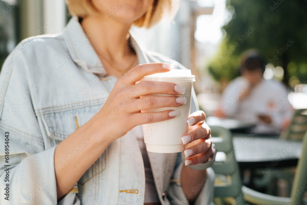 Close-up of woman in casual clothes and light manicure holding white disposable cup of coffee while sitting in street cafe on sunny summer day Close-up of woman in casual clothes and light manicure holding white disposable cup of coffee while sitting in street cafe on sunny summer day