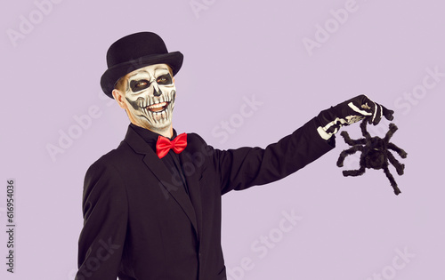 Creative Halloween. Cheerful man in Halloween costume on light lilac background poses with toy tarantula in his hand. Smiling man with skull make-up, black hat and suit with red bow tie holding spider