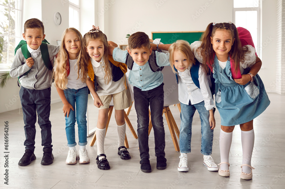 Obraz premium Group of happy children huddling in the classroom. Team of cheerful adorable friends with backpacks standing together, hugging each other, looking at the camera and smiling. Back to school concept