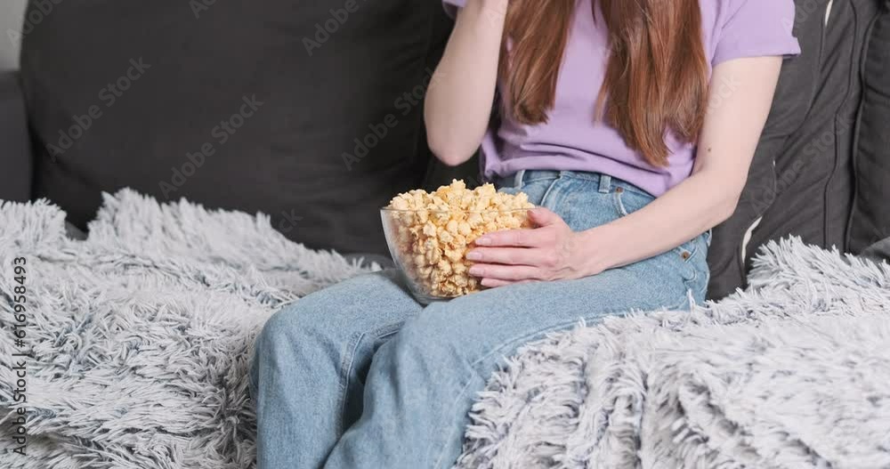 Lonely, unrecognizable woman, sitting on couch eating popcorn from glass plate while watching TV Lonely, unrecognizable woman, sitting on couch eating popcorn from glass plate while watching TV