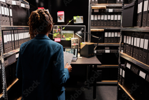 Police investigator holding crime matter file and analyzing evidence board in office late. African american woman law enforcement detective studying clues, working overtime at night