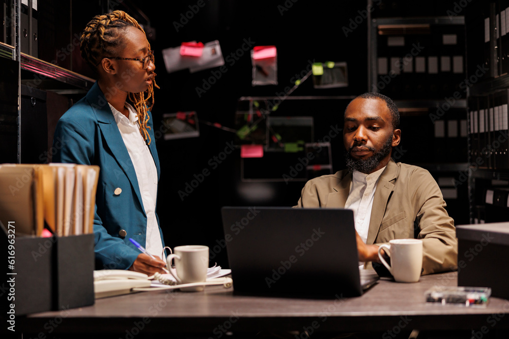 Police detective and secretary working late in office, brainstorming and analyzing records. African american woman assistant helping investigator searching crime case archival information