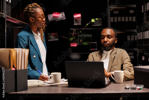 Police detective and secretary working late in office, brainstorming and analyzing records. African american woman assistant helping investigator searching crime case archival information