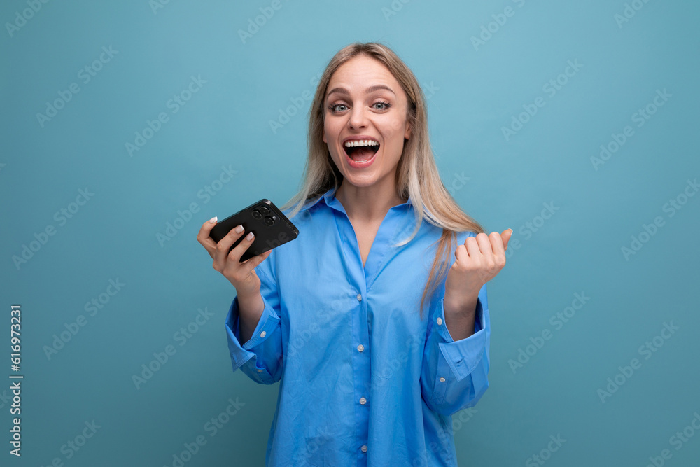 lucky charming blonde girl smiling holding smartphone in hands on isolated blue background lucky charming blonde girl smiling holding smartphone in hands on isolated blue background