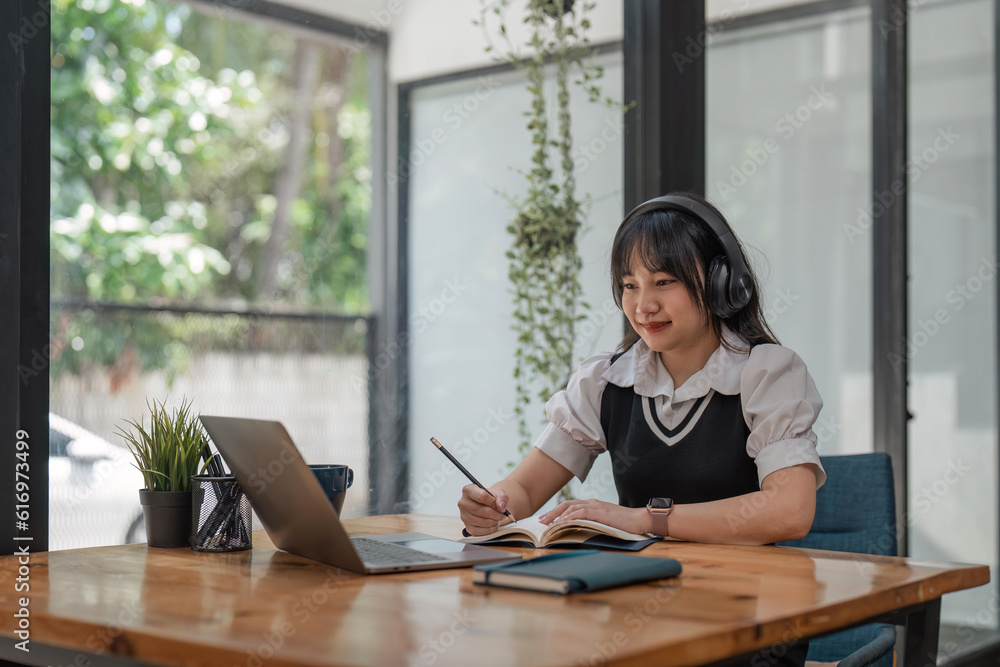 Smiling young woman in wireless headphones sitting at desk table working on laptop and writing in paper notebook Smiling young woman in wireless headphones sitting at desk table working on laptop and writing in paper notebook