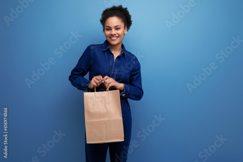 young joyful brunette latin woman dressed in blue denim overalls holding a craft bag made from ecological recycled material