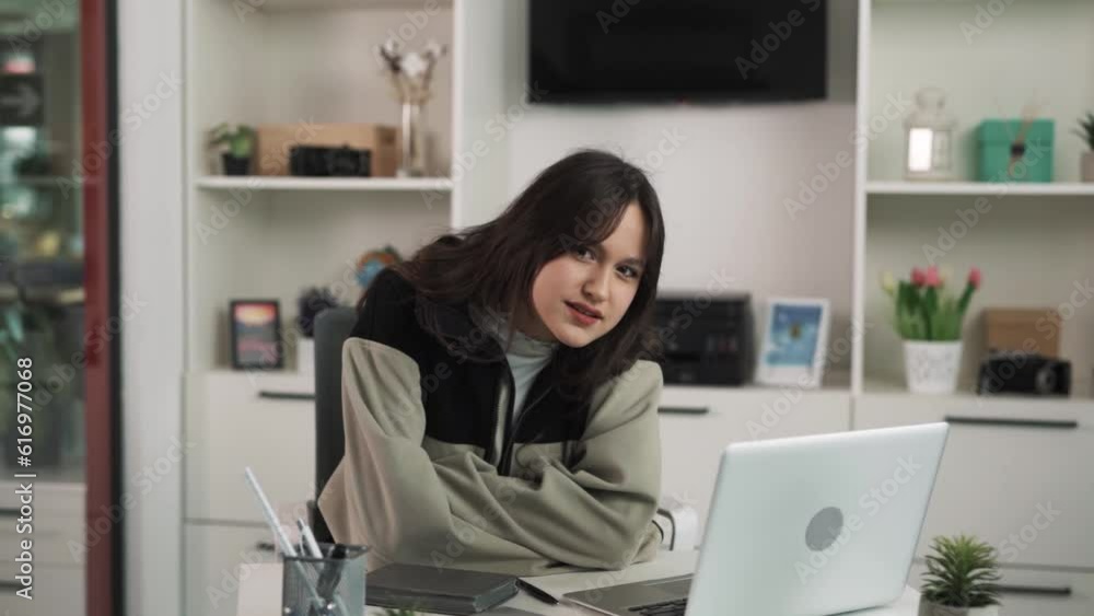 The portrait of a young dark-haired woman, arrogantly looking into the camera and saying something, sits in a bright room at a table in front of a laptop. The portrait of a young dark-haired woman, arrogantly looking into the camera and saying something, sits in a bright room at a table in front of a laptop.