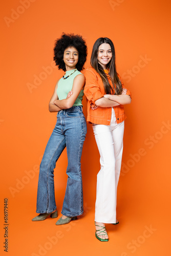 Full length of smiling and multiethnic teenage girlfriends in trendy casual outfits crossing arms and standing on orange background, teen fashionistas with impeccable style concept