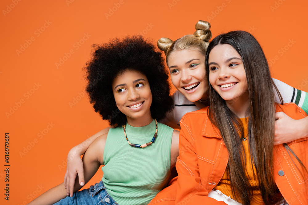 Obraz premium Cheerful blonde teenager with bold makeup hugging trendy interracial girlfriends and looking away together isolated on orange, cool and confident multicultural teenage girls