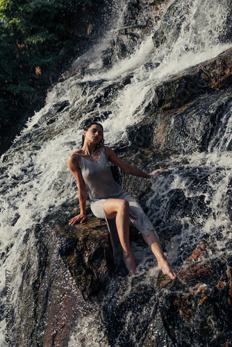 Young wet woman sitting on rock and enjoying near waterfall.