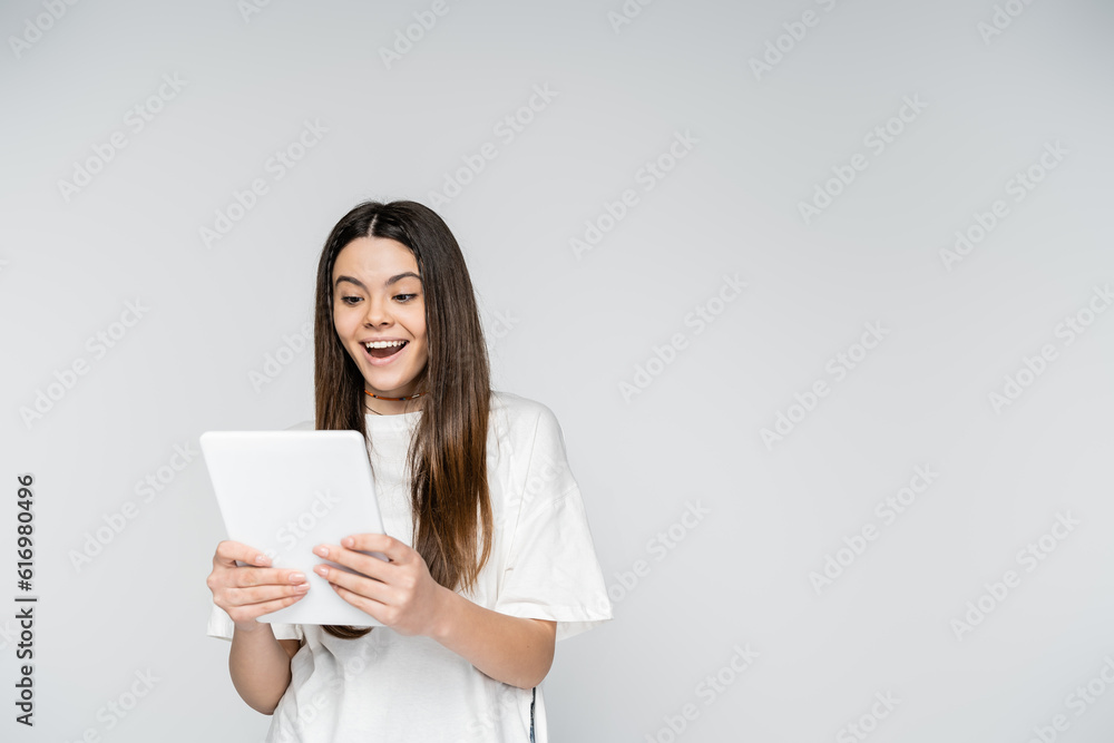 Excited brunette teenage girl in white t-shirt using digital tablet while standing isolated on grey, adolescence model and generation z concept, copy space, Touch Screen Excited brunette teenage girl in white t-shirt using digital tablet while standing isolated on grey, adolescence model and generation z concept, copy space, Touch Screen