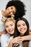 Portrait of positive multiethnic girlfriends in white t-shirts hugging brunette teen friend and looking at camera isolated on grey, energetic teenage friends spending time, friendship