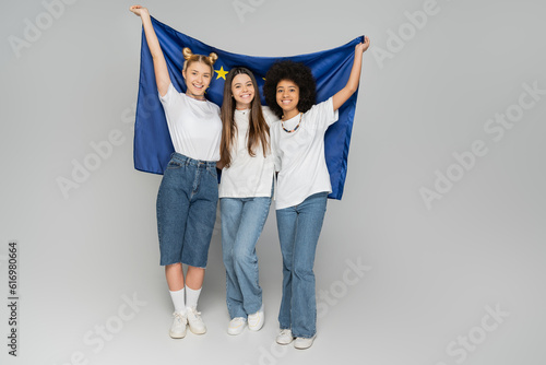 Full length of cheerful interracial teen girlfriends in white t-shirts holding European flag together and standing on grey background, energetic teenage friends spending time