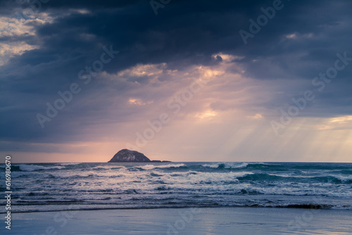 Sunset over Muriwai beach. Sun beams breaking through heavy clouds as waves splashing against the rocks. Auckland, New Zealand